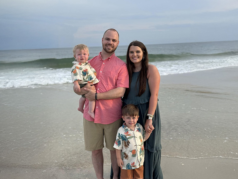 A mother, father and two sons standing in front of the ocean smiling.